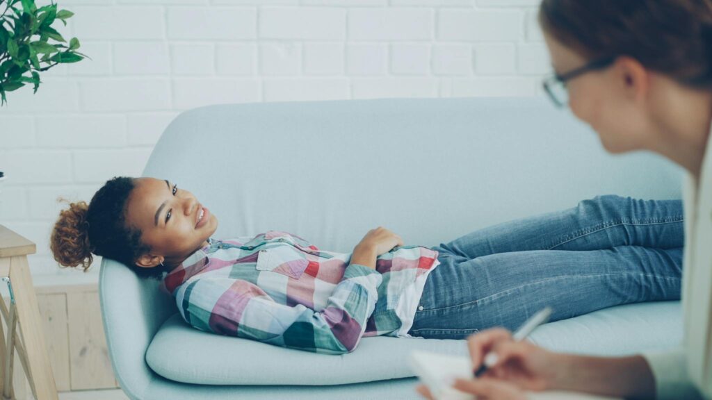 A woman in therapy, smiling while lying on a couch, with a counselor taking notes.