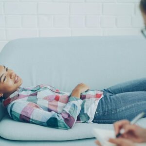 A woman in therapy, smiling while lying on a couch, with a counselor taking notes.